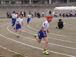 カーブのあるトラックを赤い帽子をかぶった児童が先頭を走っていて、勢いよく腕を振りながら前傾姿勢で進みほかの3人もその後ろを追っているかけっこでの写真