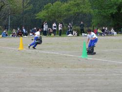 地面には緑色と黄色のコーンが立てられており、それを目印にして2人の児童が運動場で大きなビニール袋や袋状のものに下半身を入れ、ジャンプしながら進んでいる様子の写真