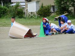 広い運動場の一角で緑色のコーンが置かれており、その横に児童がしゃがんで順番を待ち、その前の段ボールで作られた大きな筒のような物体の中に1人の児童が入っている競技の写真