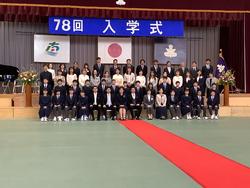 国旗や校章の上に「78回 入学式」の横断幕が設置された体育館のステージ前で、階段状に3列に並んだ生徒や保護者、教職員を斜め左方向から撮影した集合写真