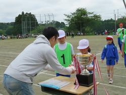 手前の大きな優勝カップが置かれた長方形の机の前に立っている関係者の男性が前に立った白い帽子の女子児童と握手をして賞状を渡している様子を写した写真