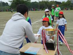 手前の大きな優勝カップが置かれた長方形の机の前に立っている関係者の男性が前に立った赤い帽子の女子児童と握手をして賞状を渡している様子を写した写真