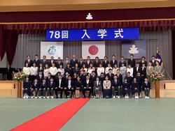 国旗や校章の上に「78回 入学式」の横断幕が設置された体育館のステージ前で、階段状に3列に並んだ生徒や保護者、教職員を斜め右方向から撮影した集合写真