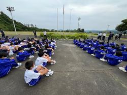 体操服姿の生徒たちが屋外の広場に集まり、整列して座り避難訓練が行われている写真