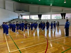 体育館の中央で青いジャージを着た生徒たちが数列に分かれて整列し、前方に立った男性と向かい合っている写真