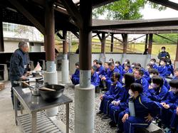 屋根付きの屋外炊事場で青いジャージを着た多くの生徒たちが並んで座り、前に立つ年配の男性が鍋や調理器具を前にして説明しているのを真剣に聞いている写真
