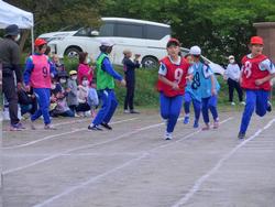 赤いビブスを着た2人の児童が前方に向かって走り、バトンを手渡して次の走者が走り出す瞬間をとらえた写真