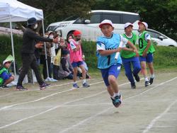 運動会でリレー競技が行われ、青いビブスを着た男子児童が右手にバトンを持って一生懸命に走っており、後方からはピンクや緑のビブスを着た他の選手たちが追いかけている写真