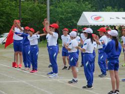 運動会の閉会式で、赤い帽子の児童たちが手を挙げ、白い帽子の児童たちが拍手をしている様子の写真