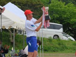 赤い帽子をかぶった男子児童が、両手で大きな優勝カップを高く掲げている写真