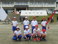 運動会の閉会後、白い帽子と赤い帽子をかぶった児童たち8人が、白組と赤組の旗や優勝トロフィーを持って運動場の中央に整列し、校舎を背景に記念撮影をしている集合写真の写真