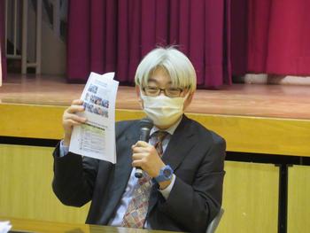 マイクを持った1人の男性が資料をみせながら説明をしている写真