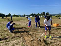 青空の下で運動着姿の生徒たちが、草地にくわやレーキを使って除草作業をしている写真