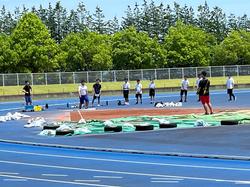 青い陸上トラックの内側で数人の生徒が整列し、中央では赤い短パンを履いた人物が指導するように立っている様子を写した写真