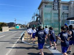 学校の横の道路沿いを、青いハーフパンツを履いた生徒たちが列になって走っている様子を後ろから写した写真