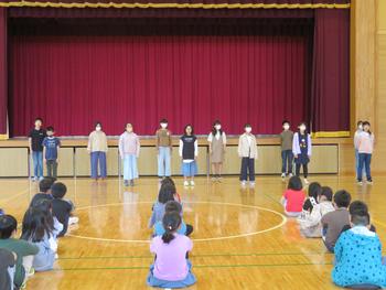 体育館の舞台前に数名の児童が並んで立ち、他の児童は間隔をあけて整列しながら床に座っている様子を写した写真