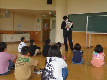 床に座った子供たちが前方の男性教師の読み聞かせに耳を傾けている写真