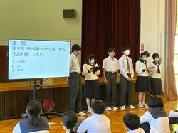 生徒たちが座る中、前方に置かれた資料が映ったスクリーンの横に、制服を着た6人の男女の生徒たちが立ち、発表をしている写真