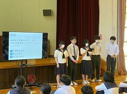 生徒たちが座る中、前方に置かれた資料が映ったスクリーンの横に、制服を着た5人の男女の生徒たちが立ち、発表をしている写真