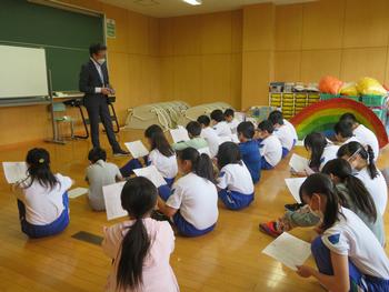体操服姿の児童達が両手で紙を持ち、床に座りながら前方の男性教師の話を聞いている写真
