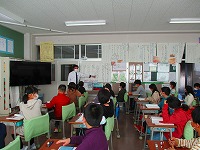 先生が教室の入口付近で掲示物を指し話をしているのを首を右に向け話を聞いている児童の写真