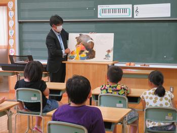 男性教員が教室の黒板の前で絵本の読み聞かせを行い、児童たちが着席しながら聞いている様子を写した写真
