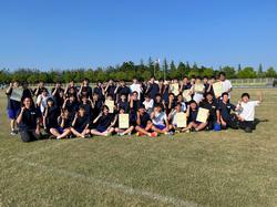 広々とした芝生のグラウンドで、大勢の生徒たちが集まり、賞状を掲げたり、ポーズをとって写した集合写真