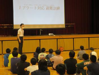 体育館の舞台に設置された大きなスクリーンに「Jアラート対応避難訓練」と映し出され、その前に立った男性の話を、床に座った児童たちが聞いている写真