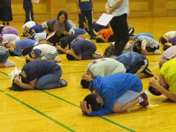 体育館内で児童たちが両手で頭を守り、小さくうずくまってダンゴムシのポーズをしている様子を写した写真
