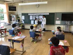 自席に座っている生徒たちが、黒板の前に立っている白衣を着た養護教諭の先生に注目している写真