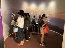 縦書きで書かれた文字が映し出された壁に囲まれた部屋で、引率の先生や子供たちが興味津々な様子で上を見上げて展示物を見ている写真