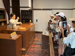 机に人形が座っている古い事務室を再現したような部屋で、子供たちが興味深そうに展示物を写真に撮っている様子の写真