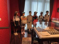 手前にはチェス盤や書類が展示されているガラスケースが設置された赤い壁の室内で、5人の子供たちが野口英世のパネルと一緒に記念撮影をしている写真