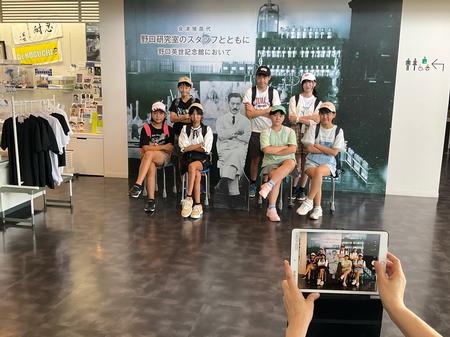 「野口研究室のスタッフとともに 野口英世記念館において」という文字が書かれ、研究室のモノクロ写真が拡大された壁の前で、足を組んだ野口英世のパネルのポーズと同じポーズをとった子供たちが2列に並び一緒に記念撮影をしている写真