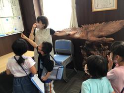 大きな木彫りの鷲が置かれている校長室で、複数の児童と女性が立って壁を見つめている中、児童の一人が掲示板を指さしている様子の写真