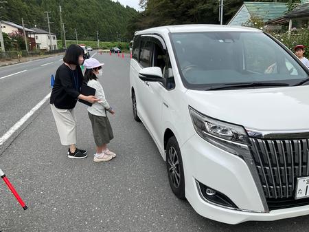 赤いコーンが並んだ道路上で、女性と一緒に立つ繭マスコットを持った女の子が、道路の端に停まっている白い車の運転手に繭マスコットを手渡そうとしている写真