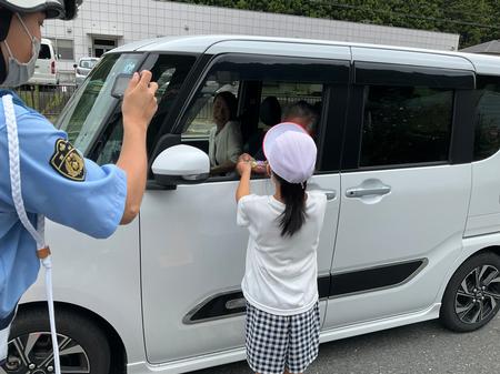 白い帽子を被った女の子が、車の助手席に乗っている人に繭マスコットを手渡している様子を、制服姿の女性警察官がカメラで撮影している写真