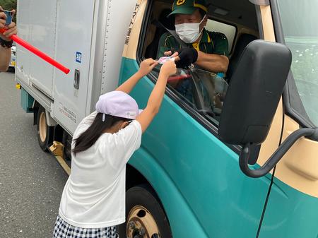 白いトラックの運転席に座っているマスクを着用している運送会社の運転手の男性に、白い帽子をかぶっている女の子が繭マスコットを渡している写真