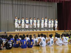 体育館のステージ上で野球のユニフォームを着た複数の選手たちが整列し、その前に他スポーツのユニフォームを着た生徒たちが座って見ている様子の写真