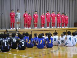 体育館のステージ上で赤いユニフォームを着た複数の選手が整列し、その前に青と白のユニフォームを着た生徒たちが座って見ている様子の写真