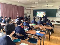 教室で生徒たちが前方の黒板前に置かれたモニターを見ながら、モニター横に立った男性の講義を受けている写真