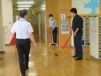 靴箱の前でさすまたやバットを持った男性職員たちが訓練を行っている写真