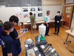 壁や展示棚に展示品が置かれた会場で、数名の来場者と2人の女子生徒が展示を見ている写真