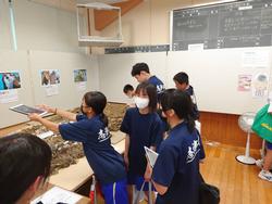 展覧会の会場で複数の生徒たちが展示物を熱心に見学しており、一人の生徒はタブレット端末を使い展示物を撮影している様子の写真