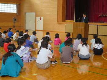 床に座った子供たちが演台に立つ男性の話を聞いている様子を写した写真