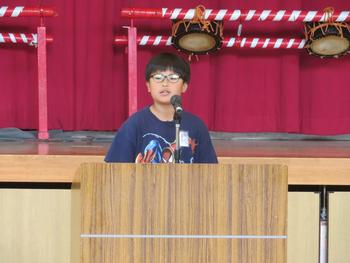 演台に立ちマイクを使って話をしているメガネをかけた男の子の写真