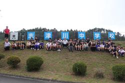 斜面上にあるフェンスに設置された「志津川中学校」の看板を背に、生徒と先生が並んで座っている写真