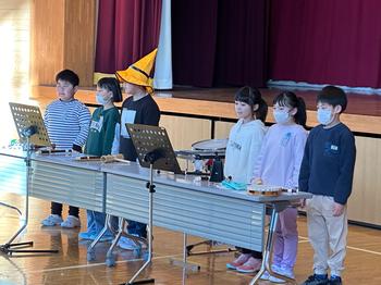 舞台の前に置いた長机の前に、3人ずつに分かれて立った6人の児童たちのうち中央の一人が黄色のとんがり帽子をかぶり、全員で合唱をしている様子の写真