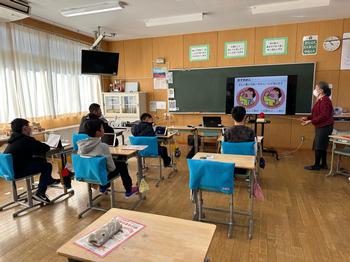 教室で小学生たちが机に座り、スクリーンに表示された教材を使って先生が授業を行っている写真