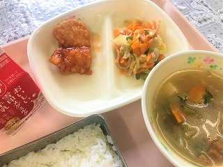 ピンクのトレーに給食が左から時計回りに牛乳、チキン南蛮、キャベツとピーマンのソテー、油麩のみそ汁、ごはんと並んでいる写真